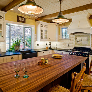 This is an example of a mid-sized beach style l-shaped eat-in kitchen in Santa Barbara with a farmhouse sink, beaded inset cabinets, white cabinets, wood benchtops, white splashback, subway tile splashback, black appliances, porcelain floors and with island.