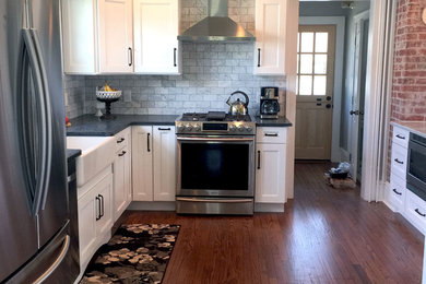 Mid-sized transitional u-shaped medium tone wood floor eat-in kitchen photo in New York with a farmhouse sink, flat-panel cabinets, white cabinets, marble countertops, gray backsplash, subway tile backsplash, stainless steel appliances and an island