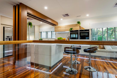 Große Moderne Wohnküche in L-Form mit Unterbauwaschbecken, flächenbündigen Schrankfronten, beigen Schränken, Quarzwerkstein-Arbeitsplatte, Küchenrückwand in Weiß, Rückwand aus Stein, schwarzen Elektrogeräten, braunem Holzboden, Kücheninsel, buntem Boden und gelber Arbeitsplatte in Brisbane