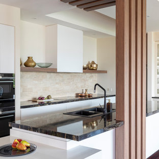 Inspiration for a large contemporary galley open plan kitchen in Brisbane with a drop-in sink, flat-panel cabinets, white cabinets, granite benchtops, beige splashback, stone tile splashback, black appliances, medium hardwood floors, brown floor, black benchtop and a peninsula.
