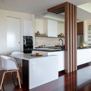 This is an example of a contemporary l-shaped kitchen in Brisbane with flat-panel cabinets, white cabinets, beige splashback, black appliances, dark hardwood floors, with island, brown floor and black benchtop.