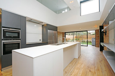 This is an example of a large contemporary galley kitchen/diner in London with a built-in sink, flat-panel cabinets, white cabinets, composite countertops, white splashback, glass sheet splashback, integrated appliances, light hardwood flooring, an island, yellow floors and white worktops.