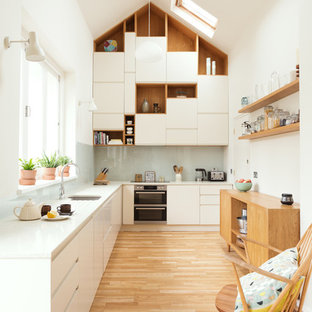 Cette image montre une cuisine design en L de taille moyenne avec un placard à porte plane, des portes de placard blanches, un plan de travail en verre recyclé, une crédence bleue, une crédence en feuille de verre, aucun îlot, un plan de travail blanc, un évier encastré, un sol en bois clair et un sol beige.