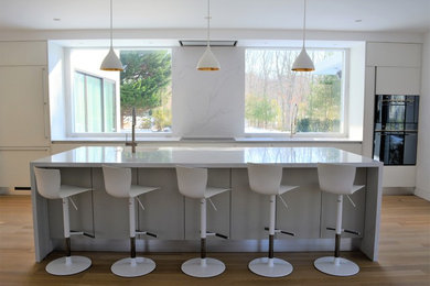 This is an example of a large contemporary single-wall enclosed kitchen in New York with an integrated sink, flat-panel cabinets, white cabinets, marble worktops, white splashback, marble splashback, black appliances, medium hardwood flooring, an island, brown floors and white worktops.