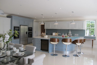 Große Moderne Wohnküche mit Schrankfronten mit vertiefter Füllung, weißen Schränken, Mineralwerkstoff-Arbeitsplatte, Küchenrückwand in Blau, Glasrückwand, Küchengeräten aus Edelstahl und Kücheninsel in Surrey