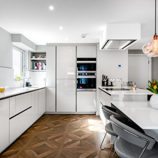 Contemporary u-shaped kitchen in London with a submerged sink, flat-panel cabinets, white cabinets, stainless steel appliances, medium hardwood flooring, a breakfast bar, brown floors and white worktops.