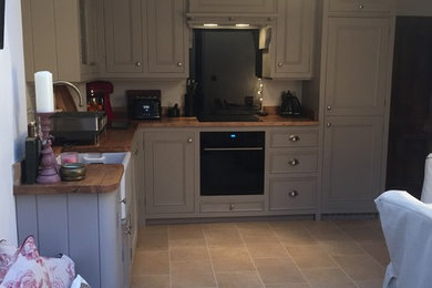 Photo of a small traditional l-shaped kitchen/diner in Other with a belfast sink, shaker cabinets, grey cabinets, wood worktops, black splashback, glass sheet splashback, black appliances, laminate floors and no island.