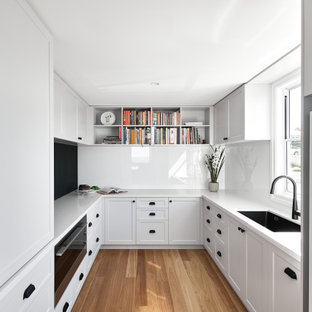 Photo of a mid-sized beach style u-shaped kitchen in Brisbane with an undermount sink, shaker cabinets, white cabinets, white splashback, black appliances, with island, brown floor and beige benchtop.