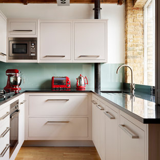 Immagine di una piccola cucina ad U design con ante lisce, ante bianche, paraspruzzi verde, paraspruzzi con lastra di vetro, elettrodomestici in acciaio inossidabile, parquet chiaro e nessuna isola