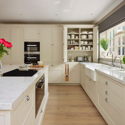 Transitional light wood floor kitchen photo in Oxfordshire with a farmhouse sink, flat-panel cabinets, beige cabinets, black appliances and an island