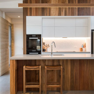 Photo of a contemporary galley kitchen in Other with a double-bowl sink, flat-panel cabinets, white cabinets, white splashback, black appliances, concrete floors, with island, grey floor and grey benchtop.