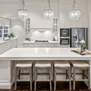 Large beach style l-shaped open plan kitchen in Brisbane with shaker cabinets, white cabinets, quartz benchtops, white splashback, porcelain splashback, stainless steel appliances, dark hardwood floors, with island, brown floor and white benchtop.