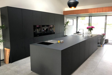 Photo of a large contemporary single-wall open plan kitchen in Cardiff with a double-bowl sink, flat-panel cabinets, black cabinets, composite countertops, black appliances, porcelain flooring, an island, beige floors and black worktops.