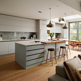 Inspiration for a large contemporary galley open plan kitchen in London with flat-panel cabinets, composite countertops, grey splashback, stainless steel appliances, an island, white worktops, a submerged sink, grey cabinets, glass sheet splashback, medium hardwood flooring and brown floors.