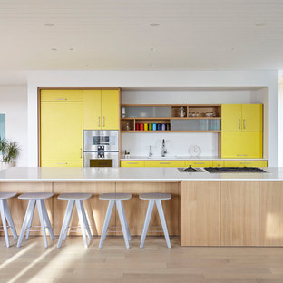 Idée de décoration pour une cuisine ouverte parallèle design de taille moyenne avec un placard à porte plane, des portes de placard jaunes, une crédence blanche, un électroménager en acier inoxydable, un sol en bois clair, un îlot central, un sol beige, un plan de travail blanc, un évier encastré et un plan de travail en surface solide.