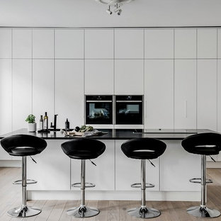 Photo of a scandinavian kitchen in Melbourne with flat-panel cabinets, white cabinets, black appliances, light hardwood floors, with island, beige floor and black benchtop.