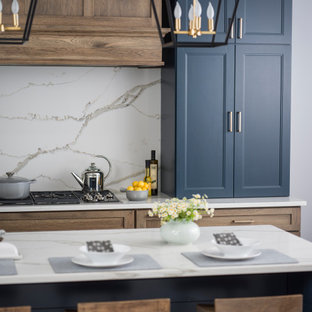 Diseño de cocina campestre, de tamaño medio, abierta, con fregadero sobremueble, armarios con paneles lisos, puertas de armario azules, encimera de cuarzo compacto, salpicadero blanco, salpicadero de losas de piedra, electrodomésticos de acero inoxidable, suelo de madera en tonos medios, una isla, suelo marrón y encimeras blancas