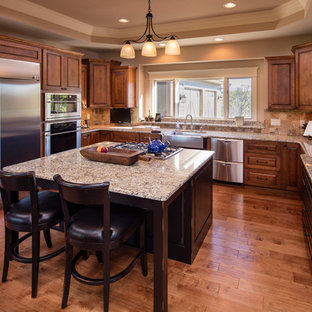 Design ideas for a large traditional u-shaped eat-in kitchen in Chicago with a farmhouse sink, raised-panel cabinets, medium wood cabinets, granite benchtops, brown splashback, slate splashback, stainless steel appliances, medium hardwood floors, with island and brown floor.