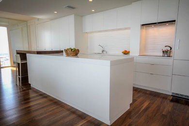 Mid-sized minimalist galley medium tone wood floor open concept kitchen photo in Tampa with an undermount sink, flat-panel cabinets, white cabinets, solid surface countertops, white backsplash, ceramic backsplash, paneled appliances and an island