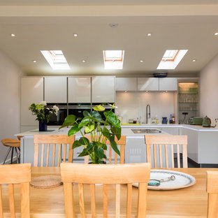 This is an example of a large contemporary single-wall open plan kitchen in Hampshire with an integrated sink, flat-panel cabinets, white cabinets, solid surface benchtops, white splashback, glass sheet splashback, stainless steel appliances, ceramic floors and with island.