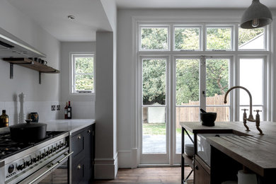 This is an example of a medium sized victorian single-wall enclosed kitchen in London with a belfast sink, shaker cabinets, grey cabinets, wood worktops, white splashback, porcelain splashback, stainless steel appliances, medium hardwood flooring, an island, brown floors and brown worktops.