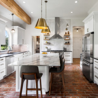 Modelo de cocina comedor en U, grande, con fregadero bajoencimera, armarios con paneles empotrados, puertas de armario blancas, encimera de cuarcita, salpicadero blanco, salpicadero de azulejos tipo metro, electrodomésticos de acero inoxidable, suelo de ladrillo, una isla, suelo marrón y encimeras grises