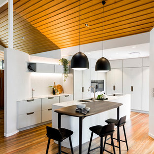 Photo of a mid-sized contemporary l-shaped eat-in kitchen in Brisbane with an undermount sink, flat-panel cabinets, white cabinets, white splashback, black appliances, medium hardwood floors, with island, brown floor and white benchtop.