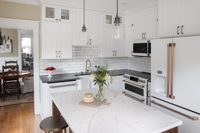 Photo of a small classic l-shaped enclosed kitchen in Other with white cabinets, engineered stone countertops, white splashback, metro tiled splashback, white appliances, medium hardwood flooring, an island, brown floors, white worktops, a belfast sink and shaker cabinets.