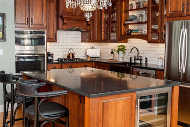 Large elegant l-shaped medium tone wood floor and brown floor eat-in kitchen photo in Providence with a farmhouse sink, glass-front cabinets, medium tone wood cabinets, solid surface countertops, white backsplash, subway tile backsplash, stainless steel appliances, an island and black countertops