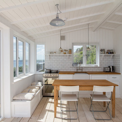 Example of a mid-sized beach style single-wall light wood floor eat-in kitchen design in Cornwall with a farmhouse sink, white cabinets, white backsplash, subway tile backsplash, stainless steel appliances, shaker cabinets and wood countertops