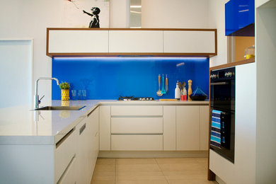 クライストチャーチにある中くらいなコンテンポラリースタイルのおしゃれなキッチン (シングルシンク、フラットパネル扉のキャビネット、白いキャビネット、ラミネートカウンター、青いキッチンパネル、ガラス板のキッチンパネル、黒い調理設備、磁器タイルの床、ベージュの床、白いキッチンカウンター) の写真