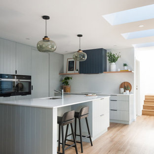 Small beach style l-shaped kitchen in Central Coast with an undermount sink, blue cabinets, quartz benchtops, engineered quartz splashback, light hardwood floors, with island, beige floor, flat-panel cabinets, white splashback, black appliances and white benchtop.