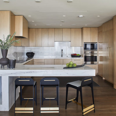 Example of a trendy u-shaped dark wood floor and brown floor kitchen design in Los Angeles with an undermount sink, flat-panel cabinets, white cabinets, white backsplash, stone slab backsplash, stainless steel appliances, a peninsula and white countertops