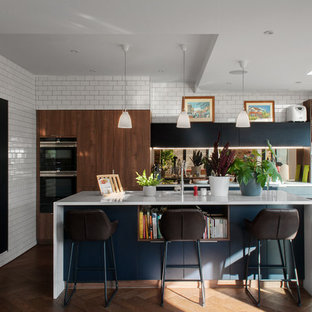 Design ideas for a medium sized contemporary l-shaped open plan kitchen in London with flat-panel cabinets, composite countertops, medium hardwood flooring, an island, white worktops, a submerged sink, medium wood cabinets, brown floors and mirror splashback.