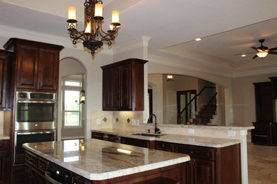 Example of a mid-sized classic l-shaped travertine floor eat-in kitchen design in Houston with an undermount sink, raised-panel cabinets, dark wood cabinets, granite countertops, beige backsplash, stone tile backsplash, stainless steel appliances and an island