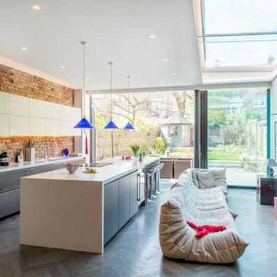 Example of a large trendy single-wall dark wood floor and gray floor open concept kitchen design in London with an integrated sink, flat-panel cabinets, gray cabinets, solid surface countertops, pink backsplash, brick backsplash, stainless steel appliances and an island