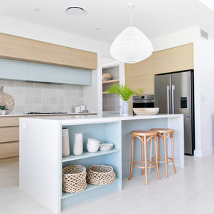Inspiration for a mid-sized beach style galley open plan kitchen in Gold Coast - Tweed with an undermount sink, flat-panel cabinets, light wood cabinets, quartz benchtops, beige splashback, ceramic splashback, stainless steel appliances, ceramic floors, with island, white floor and white benchtop.