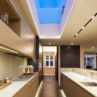 Example of a large trendy galley dark wood floor and brown floor kitchen design in London with an undermount sink, flat-panel cabinets, solid surface countertops, beige backsplash, glass tile backsplash, paneled appliances, white countertops, beige cabinets and no island