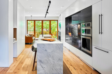 Example of a mid-sized trendy galley medium tone wood floor eat-in kitchen design in Toronto with flat-panel cabinets, white cabinets, marble countertops, black backsplash, slate backsplash, stainless steel appliances, an island and white countertops