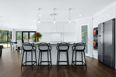 Foto på ett mellanstort funkis vit kök, med en dubbel diskho, vita skåp, bänkskiva i kvarts, vitt stänkskydd, glaspanel som stänkskydd, rostfria vitvaror, mörkt trägolv, en köksö och brunt golv