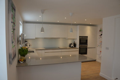 This is an example of a large contemporary u-shaped kitchen/diner in Buckinghamshire with a submerged sink, flat-panel cabinets, white cabinets, quartz worktops, white splashback, glass sheet splashback, stainless steel appliances, light hardwood flooring and a breakfast bar.