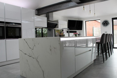 This is an example of a large contemporary kitchen/diner in Essex with glass-front cabinets, white cabinets, grey splashback, glass sheet splashback, black appliances and an island.
