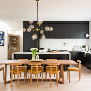 Réalisation d'une grand cuisine américaine design en L avec un évier encastré, un placard à porte plane, des portes de placard noires, un plan de travail en quartz, une crédence blanche, une crédence en quartz modifié, un électroménager en acier inoxydable, un sol en bois clair, un îlot central, un sol marron et un plan de travail blanc.