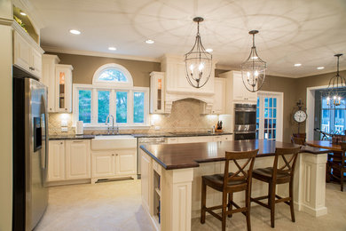 Example of a large classic l-shaped travertine floor enclosed kitchen design in Indianapolis with a farmhouse sink, raised-panel cabinets, white cabinets, granite countertops, beige backsplash, ceramic backsplash, stainless steel appliances and an island