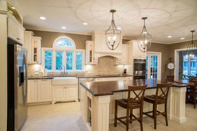 Photo of a large classic l-shaped enclosed kitchen in Indianapolis with a belfast sink, raised-panel cabinets, white cabinets, granite worktops, beige splashback, ceramic splashback, stainless steel appliances, travertine flooring and an island.