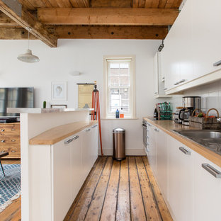 Industrial single-wall kitchen in London with a built-in sink, flat-panel cabinets, white cabinets, wood worktops, white splashback, medium hardwood flooring, an island and brown floors.