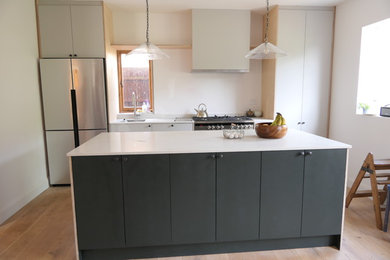 Photo of a medium sized industrial single-wall open plan kitchen in Gloucestershire with an integrated sink, flat-panel cabinets, green cabinets, engineered stone countertops, stainless steel appliances, light hardwood flooring, an island, brown floors and white worktops.