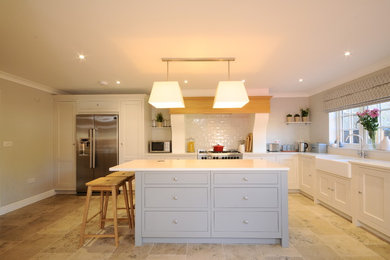 Photo of a large contemporary l-shaped kitchen/diner in Cambridgeshire with a belfast sink, flat-panel cabinets, grey cabinets, composite countertops, white splashback, ceramic splashback, stainless steel appliances, limestone flooring, an island, beige floors and white worktops.