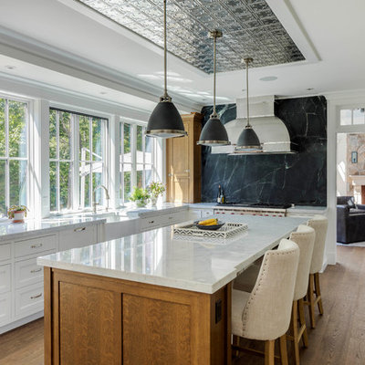 Example of a large classic l-shaped medium tone wood floor and brown floor enclosed kitchen design in Boston with a farmhouse sink, shaker cabinets, white cabinets, marble countertops, black backsplash, stone slab backsplash, stainless steel appliances, an island and gray countertops