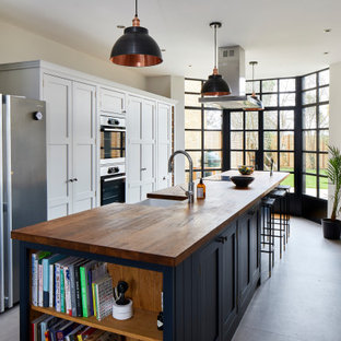 This is an example of a classic galley enclosed kitchen in London with a belfast sink, shaker cabinets, white cabinets, wood worktops, stainless steel appliances, concrete flooring, an island, grey floors and brown worktops.